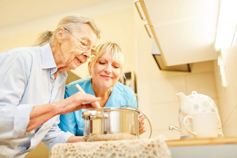 Pflegedienst Frau und Seniorin in der Küche der Seniorenwohnung beim Kochen